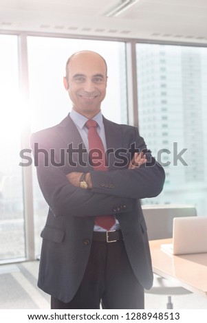 Portrait of smiling businessman standing with arms crossed in boardroom at office with lens flare in background 