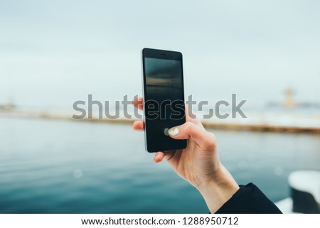 Close-up of female hand holding mobile phone photographing seascape on winter day.