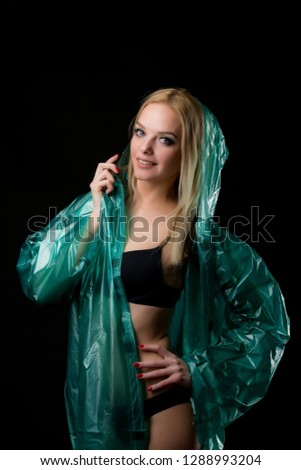 Portrait of a young blond woman in a green raincoat looking at the camera, a hood is put on her head. Shooting in a photo studio on a black background