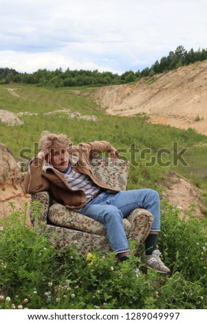young man with white heir sits in a old chair in a green field alone