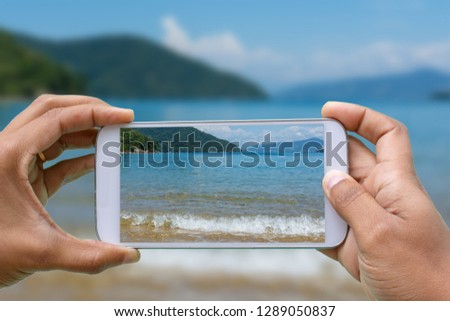 hands with smartphone photographing Praia de Abraaozinho, sea of crystalline and calm waters, surrounded by vegetation in Ilha Grande, Angra dos Reis, Rio de Janeiro