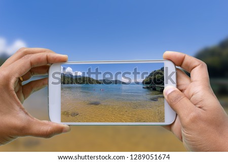 hands with smartphone photographing Praia de Abraaozinho, sea of crystalline and calm waters, surrounded by vegetation in Ilha Grande, Angra dos Reis, Rio de Janeiro