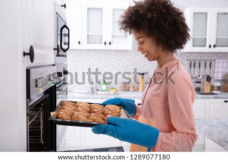 A Happy Young Woman Wearing Blue Mitt Removing Baked Croissants Tray From An Oven Royalty-Free Stock Photo #1289077180