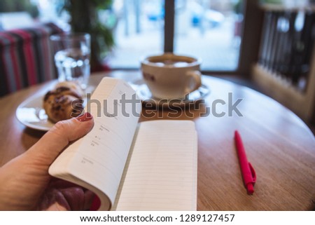 A red note and red pen is on the table in a cafe, a cup of coffee and a croissant on background. Female hand holding a notepad