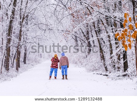 Older man and woman walking on trail in beautiful snowy park during snowfall; woman is taking pictures with her smartphone