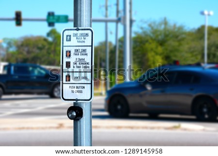 Traffic intersection pedestrian crosswalk crossing sign with signal descriptions above the request button with cars and lights in the background.