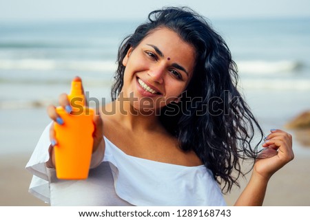 portrait of a beautiful and smiling snow-white smile indian woman black curly hair and dark skin in a white t-shirt holding bottle of sunscreen spray on beach.girl enjoying spf body paradise vacation
