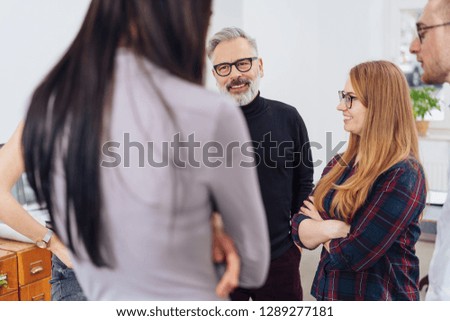 Businessman standing chatting with his team in an office in an over the shoulder view of him smiling
