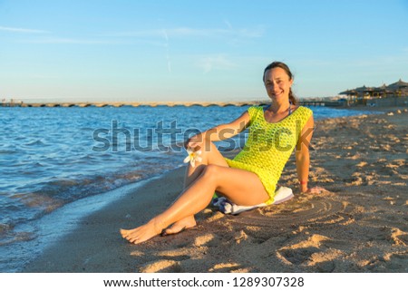 Happy young woman sitting on beach. woman sitting on sandy beach against blue sky outdoors. An attractive young woman sits on a beach with her elbow on her knee. Horizontal shot.