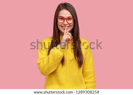 Positive woman demonstrates hush gesture, keeps fore finger over lips, dressed in casual outfit, gossips with best friend, tells secret information, models over pink background, wears yellow sweater