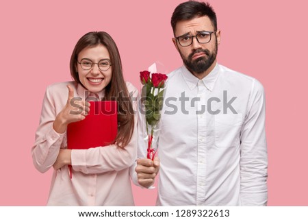Photo of attractive woman shows approval gesture, keeps thumb up, carries red notepad, puzzled unshaven man holds bouquet of roses, feels awkward, has first love. People and relations concept