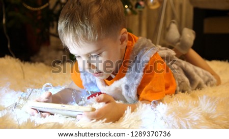 The child lies on a soft white blanket in the children's room. He is watching cartoons on the smartphone. Christmas garland. Happy childhood