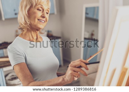 Enjoy your hobby. Pleased female person keeping smile on her face while drawing picture with pleasure