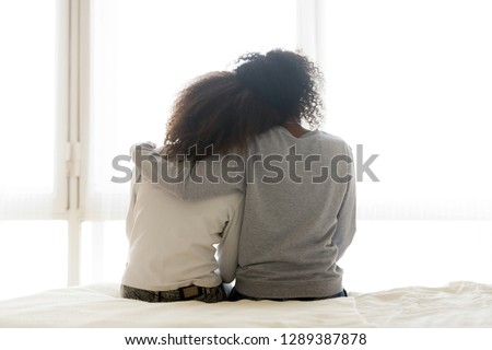 Back view of loving African American mother hug teen daughter sitting on bed, caring black mom embrace child, relaxing in bedroom looking in window, parent comfort teenager caressing at home Royalty-Free Stock Photo #1289387878
