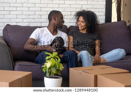 Young african american couple happy to move into new home with pet and boxes, black family tenants celebrate relocation sitting on couch with dog, homeowners renters having fun unpacking in own house Royalty-Free Stock Photo #1289448100