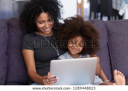 Smiling african american mother and kid daughter having fun with computer sitting on couch, happy mixed race mom with child girl using laptop, watching cartoons, making video call, doing shopping