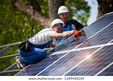 Professional technicians working with electrical screwdriver installing shiny solar photo voltaic panel to metal platform system on green tree thick foliage background.