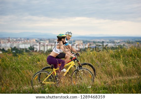 Two positive and energetic cyclists riding bicycle on trail in high grass. Beautiful and cheerful man and woman in sportswear and helmets. Behind them city and built environment. Royalty-Free Stock Photo #1289468359