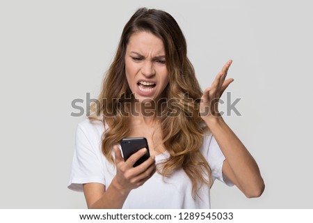 Annoyed angry young woman mad about spam message stuck phone looking at smartphone isolated on blank studio background, furious teen girl having problem with cellphone irritated by broken mobile Royalty-Free Stock Photo #1289545033