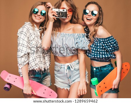 Three beautiful stylish smiling girls with colorful penny skateboards.Women in summer hipster checkered shirt clothes posing on golden background.Taking pictures on retro camera