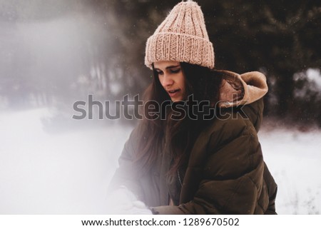Close up portrait of happy woman which wear jacket and hoodie enjoying winter moments. Outdoor photo of long-haired lady in knitted hat having fun in snowy morning on blur nature background.