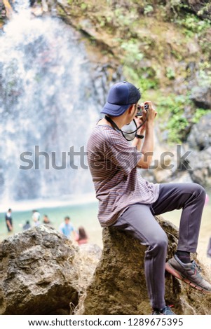Young man sit and turn back taking photo of waterfall.
