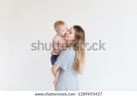 Scene of pure love and happiness. Full lenght shot of lovely young happy mother kissing her little baby over white wall in sunny bedroom at home. Mom and kid spend joyfully time together
