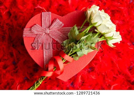 Heart shaped boxed gift with white roses bouquet, placed on red feathers background. Valentines day or Romantic date concept. Top view shot. With copy space