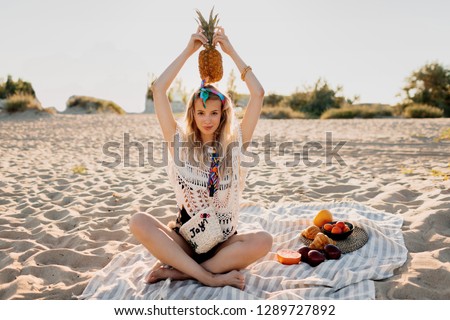 Summertime picture of lovely blond woman holding pineapple on her head and sitting on beach towel in sunny evening. Tropical mood. Boho outfit.