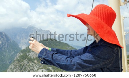 the girl in sunglasses and red hat takes a photos and video with sea, mountain and forest in sunny summer day