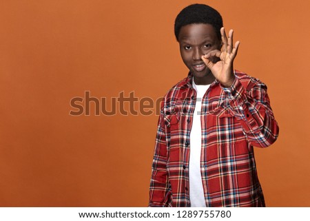 I got it. Studio shot of friendly looking dark skinned hipster guy dressed in checkered shirt smiling happily, circling thumb and fore finger, showing ok gesture as sign of agreement or approval