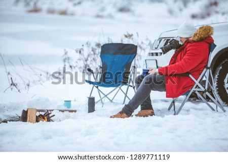 young adult man sitting in chair near winter camp fire taking picture on camera. travel concept