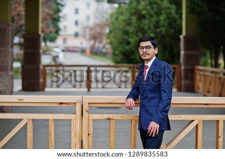Indian young man at glasses, wear on suit with red tie posed outdoor.