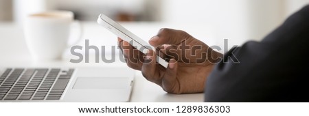 Horizontal concept photo african businessman sitting at desk near pc close up hand hold use smartphone app communicate online browse internet, banner for website header design with copy space for text
