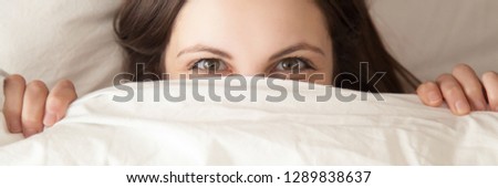 Horizontal top close up shy playful woman looking smiling eyes at camera hiding under white blanket lying on pillow in bed having fun concept, banner for website header design with copy space for text
