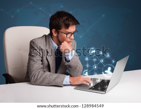 Attractive young man sitting at desk and typing on laptop with social network icons comming out