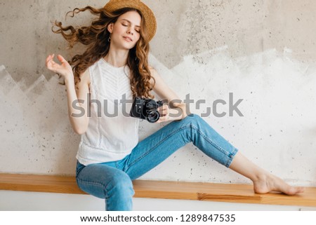 young pretty smiling woman wearing blue denim jeans and white shirt sitting against wall in straw hat holding vintage photo camera, traveler in summer outfit, fashion trend, waving long curly hair