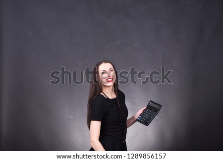 Young beautiful woman (brunette) with red lips and with a smile, long dark hair holds a calculator on a black background. Profit and expenses. Studio