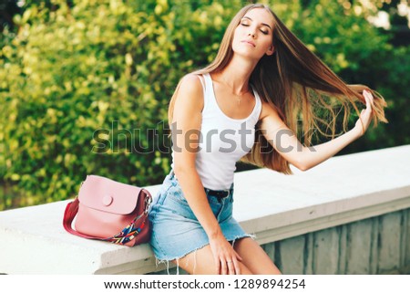 Young beautiful stylish woman in white shirt and denim skirt with pink bag.   Lovable long-haired blonde woman enjoying life and having fun at resort, happy, street style, spring summer trend.