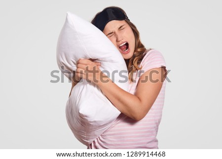 Image of attractive woman yawns as has sleepy expression, wears domestic clothes, carries soft white pillow, poses against white background. People, rest, comfort, tiredness, sleeping concept