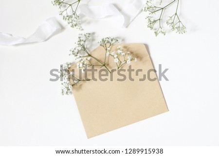 Styled stock photo. Feminine wedding desktop mockup with baby's breath Gypsophila flowers, satin ribbon and blank craft paper envelope mockup on white table background. Top view, flat lay.