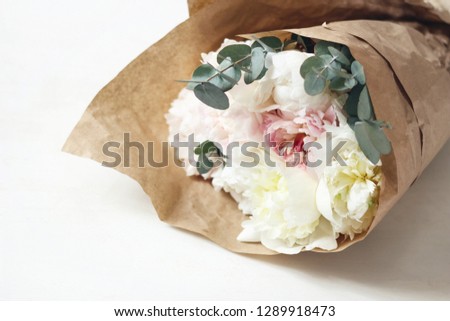 Bouquet of soft pink and white peony flowers and eucalyptus branches wrapped in brown kraft paper on white table background. Feminine style stock photo. Selective focus.
