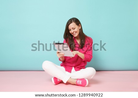 Full length portrait woman in rose shirt blouse, white pants sitting on floor hold tablet pc isolated on pink blue pastel wall background studio. Fashion lifestyle concept. Mock up copy space.