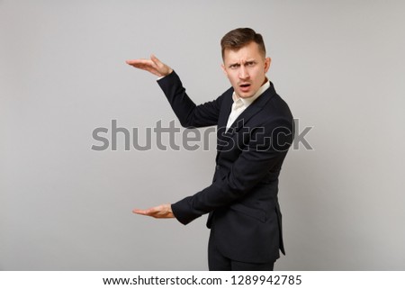 Concerned young business man in classic black suit gesturing demonstrating size with vertical copy space isolated on grey wall background. Achievement career wealth business concept. Advertising area