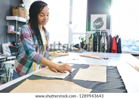Young female tailor working with sketches and textile and sewing new dress in design studio