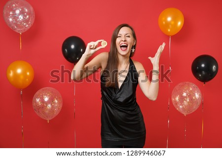 Overjoyed woman in black dress spreading hands holding bitcoin metal coin of golden color, future currency on bright red background air balloons. Happy New Year, birthday mockup holiday party concept