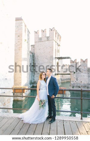 Portrait of cheerful young beautiful couple in blue clothes smiling and hugging on the background of Sirmione castle in the morning