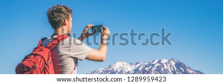 Travel tourist hiking man taking picture with phone of mountains landscape on summer hike adventure. Panorama banner with copy space on blue sky background.