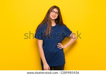 Young woman with glasses over yellow wall posing with arms at hip and smiling