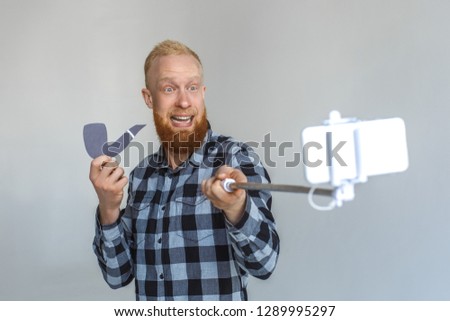 Red hair mature man standing isolated on grey wall holding monopod taking selfie photo on smartphone with paper tobacco pipe looking camera smiling cheerful